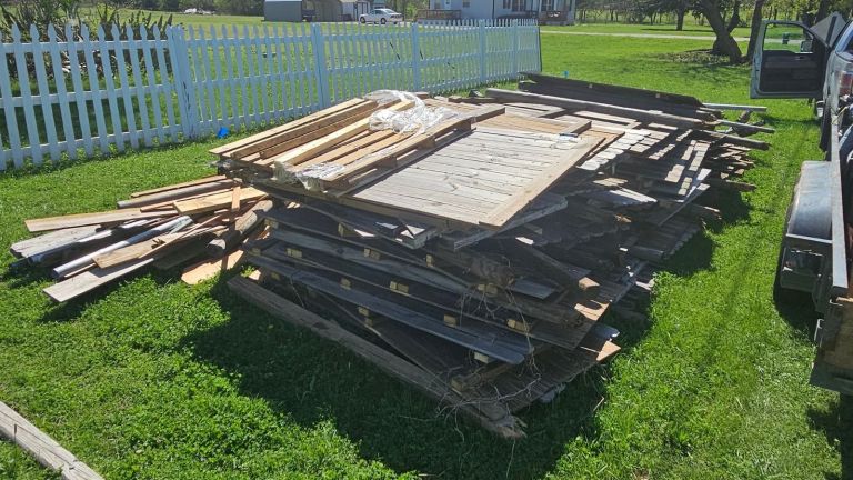 A large pile of wooden boards and planks, likely from a fence removal, lies on green grass near a white picket fence, with a trailer and vehicle partially visible on the right side.