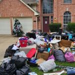 A large pile of garbage bags, boxes, and discarded items sits on the curb outside a brick house with two garage doors and trimmed bushes—an eviction cleanout in progress. A Christmas tree stands near the garage, partially decorated.