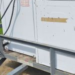 A white washing machine and dryer sit on a metal utility trailer parked on a street near green grass, awaiting appliance removal. Both appliances appear weathered, with some dirt and tape visible on the surfaces.