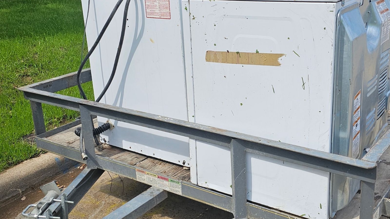 A white washing machine and dryer sit on a metal utility trailer parked on a street near green grass, awaiting appliance removal. Both appliances appear weathered, with some dirt and tape visible on the surfaces.