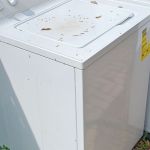 A white washing machine with dirt and leaves on the lid sits outdoors beside a dryer, near a wooden fence and brick pavement—ideal for appliance removal.