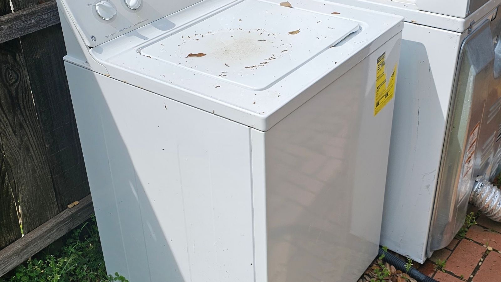 A white washing machine with dirt and leaves on the lid sits outdoors beside a dryer, near a wooden fence and brick pavement—ideal for appliance removal.