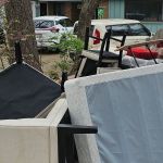 A pile of furniture, including chairs, a mattress, and sofa cushions, is stacked outside in front of a house with two parked cars and trees nearby—typical of an eviction cleanout awaiting junk removal.