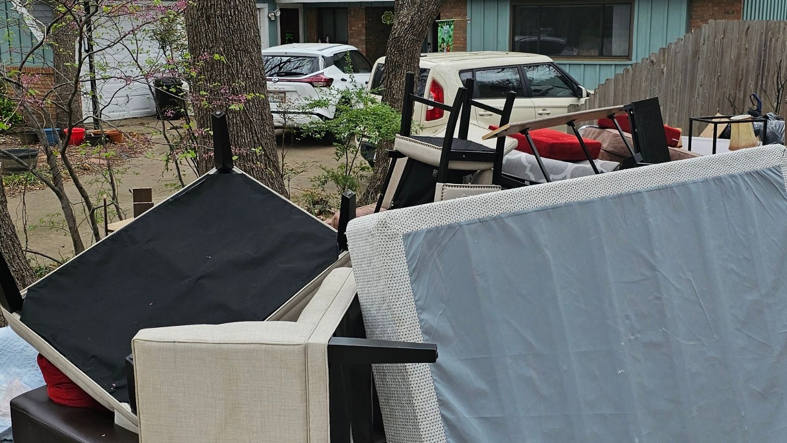 A pile of furniture, including chairs, a mattress, and sofa cushions, is stacked outside in front of a house with two parked cars and trees nearby—typical of an eviction cleanout awaiting junk removal.
