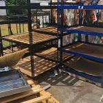 Metal and wooden shelves stand empty outdoors, surrounded by broken down cardboard boxes and discarded packaging—remnants awaiting scrap metal removal in Dallas, TX by Arlington Junk Removal Service under the bright sunlight.