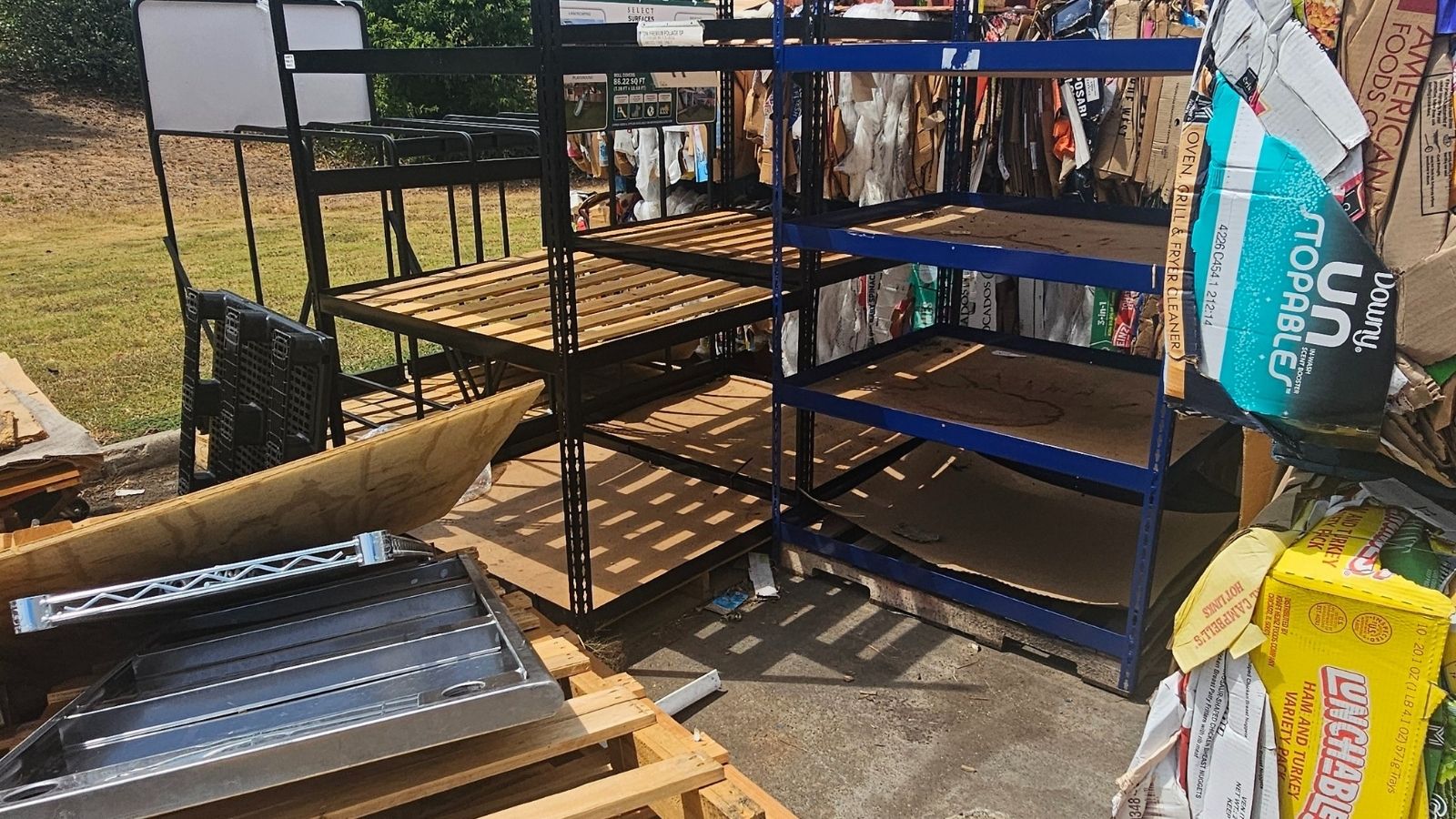 Metal and wooden shelves stand empty outdoors, surrounded by broken down cardboard boxes and discarded packaging—remnants awaiting scrap metal removal in Dallas, TX by Arlington Junk Removal Service under the bright sunlight.