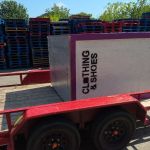 A large white donation bin labeled CLOTHING & SHOES sits tilted on a red trailer, ready for donation box removal in Dallas, with stacks of colorful pallets and trees visible in the background.