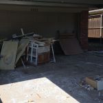 A pile of discarded furniture, wood, and construction debris sits in front of a closed garage door at house number 2321. Pieces of a broken drawer and other trash are scattered across the driveway, awaiting construction debris removal.