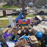 A large pile of household items, boxes, bags, and furniture sits on a front lawn near a sidewalk—ready for eviction cleanout. A white pickup truck with a trailer is parked on the street nearby under a leafless tree.