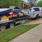 A pickup truck with an open door is parked on a residential street, towing a trailer loaded with furniture, mattresses, and household items—possibly part of a junk removal service or cleanout.