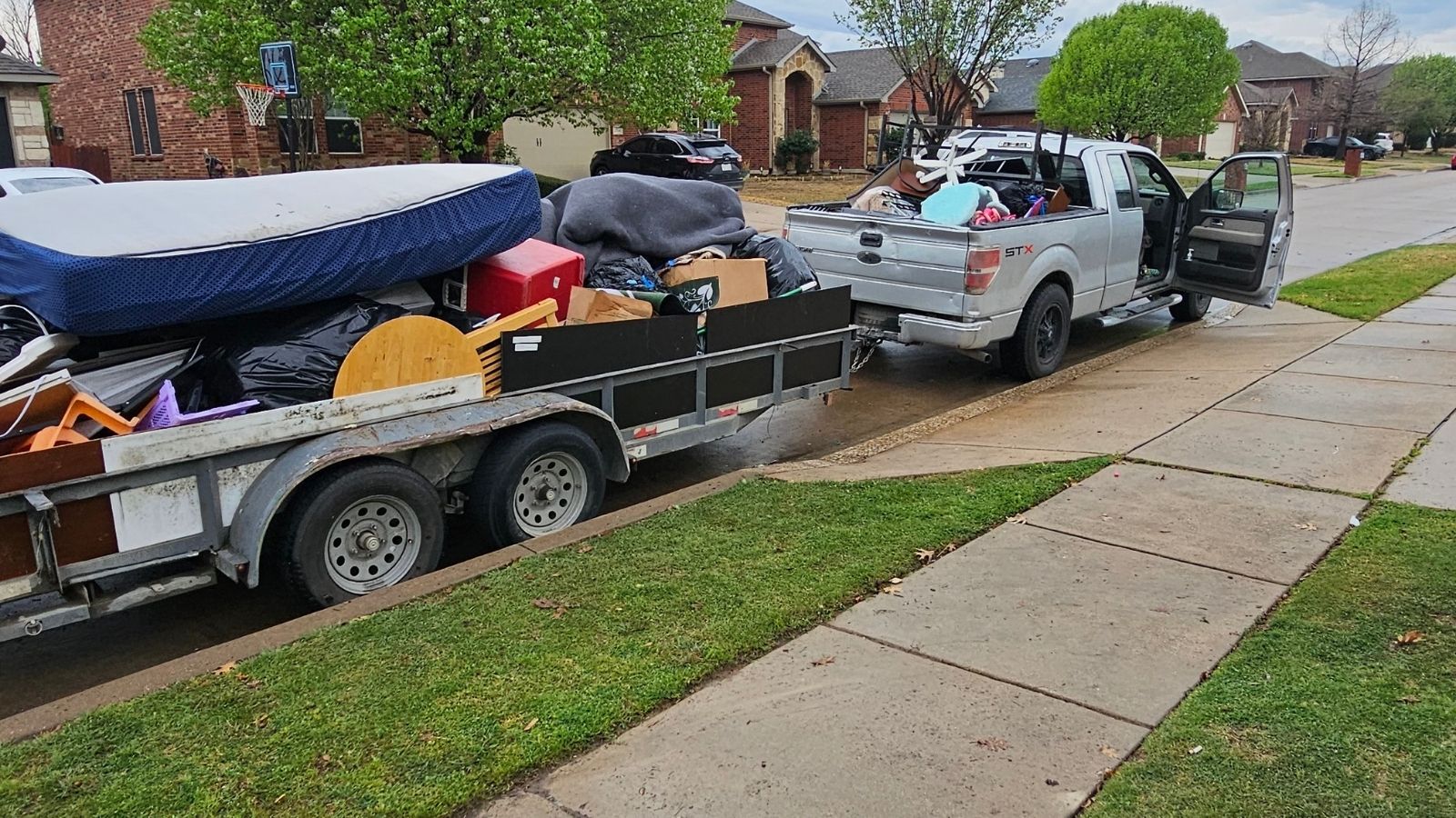 A pickup truck with an open door is parked on a residential street, towing a trailer loaded with furniture, mattresses, and household items—possibly part of a junk removal service or cleanout.