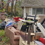 A large pile of discarded furniture, bags, and household items from an eviction cleanup is stacked on the front lawn of a house with beige siding and white trim, next to a tree with budding pink flowers.