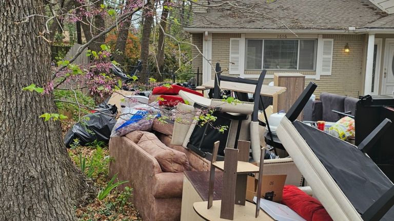 A large pile of discarded furniture, bags, and household items from an eviction cleanup is stacked on the front lawn of a house with beige siding and white trim, next to a tree with budding pink flowers.