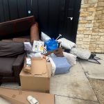 A pile of discarded furniture and household items, including a brown sofa, cardboard boxes, and bags, sits beside a stone building and black doors on a concrete sidewalk awaiting trash removal.