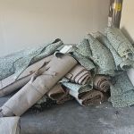 Rolled-up pieces of old carpet and underpad from a recent carpet removal are stacked in a pile on a concrete floor near a white door, with some debris scattered around the room or garage.