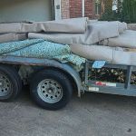 A trailer loaded with rolled-up carpet pieces from a recent carpet removal and plant debris is parked on a driveway in front of a garage and a brick house.