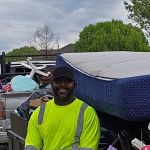 A smiling man in a neon yellow safety shirt and black cap stands next to a pickup truck filled with household items, showcasing his junk removal service on a tree-lined residential street.