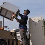 A person lifts a small refrigerator onto a trailer filled with old appliances and furniture, next to a white fridge with magnets—an efficient junk removal service at work outdoors on a sunny day.