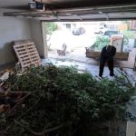 Two people tackle a garage cleanout, clearing away a large pile of branches and yard debris. The garage door is open, with pallets and boxes stacked nearby as daylight shines in from outside.