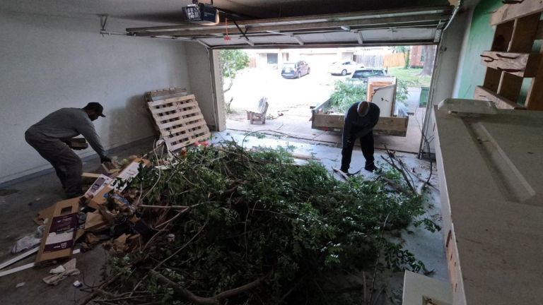 Two people tackle a garage cleanout, clearing away a large pile of branches and yard debris. The garage door is open, with pallets and boxes stacked nearby as daylight shines in from outside.