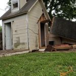 A small shed with severe damage to one side, its wall and roof partially collapsed, stands ready for demolition. A person in a neon yellow shirt stands nearby on grassy ground, facing the structure prepared for shed removal.
