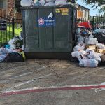 An overflowing dumpster with large piles of garbage bags and discarded items scattered on the ground around it awaits trash removal in a residential area next to a fenced-in grassy hill.