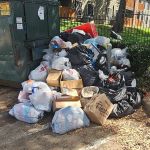 A large pile of trash bags, cardboard boxes, and loose garbage is stacked next to an overflowing dumpster in a parking lot near an apartment complex, highlighting the urgent need for trash removal near the fenced grassy area.