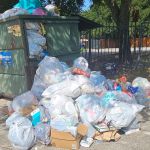 An overflowing dumpster with numerous plastic bags and loose trash piled up on the ground around it, near a fence and some trees on a sunny day, highlighting the urgent need for trash removal.
