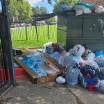 Piles of trash bags, clothes, and cardboard boxes are scattered on the ground next to an overflowing dumpster near a gated fence, highlighting the need for prompt trash removal with grass and buildings visible in the background.