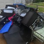 A pile of assorted household items, storage bins, trash bags, black foam mats, and a metal bench with a green cushion await junk removal on a wet driveway near the garage door, next to the sidewalk and grass.