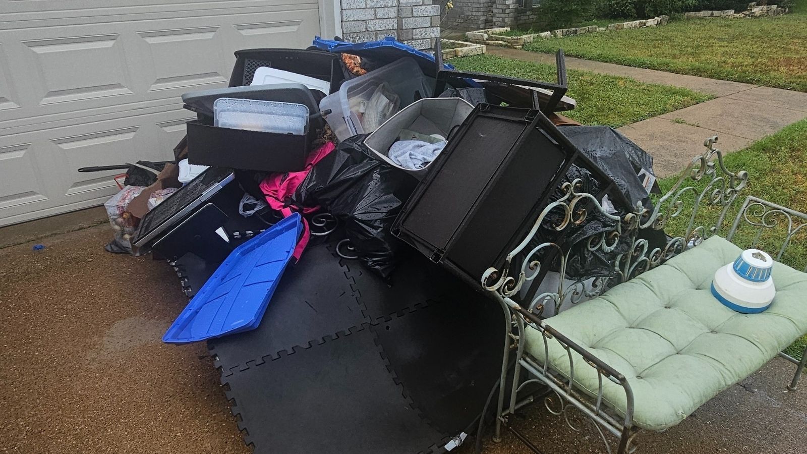 A pile of assorted household items, storage bins, trash bags, black foam mats, and a metal bench with a green cushion await junk removal on a wet driveway near the garage door, next to the sidewalk and grass.