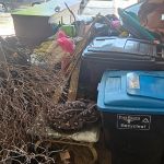 A cluttered garage filled with recycling bins, branches, trash bags, and miscellaneous items—including a blanket and toys—all piled together in a messy heap, awaiting a thorough garage cleanout.