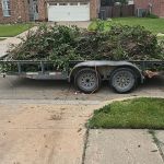 A flatbed trailer attached to a pickup truck is filled with a large pile of branches and yard debris, ready for yard debris removal, parked on a residential street near a driveway and brick mailbox.