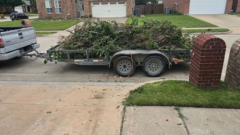 A flatbed trailer attached to a pickup truck is filled with a large pile of branches and yard debris, ready for yard debris removal, parked on a residential street near a driveway and brick mailbox.