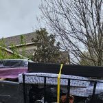 A metal trailer loaded with mattresses, furniture, and other household items for mattress removal is secured with yellow straps and parked outside a brick house on a cloudy day, with bare trees and a residential street in the background.