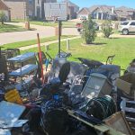 A large pile of assorted household items, furniture, and black trash bags sits on a driveway after an eviction cleanout in front of a suburban house on a sunny day, with other houses and cars visible in the background.