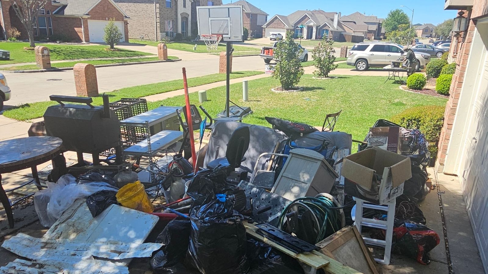 A large pile of assorted household items, furniture, and black trash bags sits on a driveway after an eviction cleanout in front of a suburban house on a sunny day, with other houses and cars visible in the background.