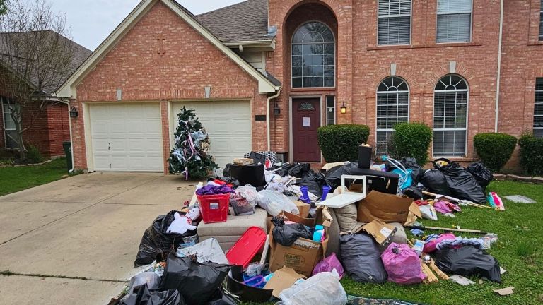 A large pile of garbage bags, boxes, and discarded items—including a Christmas tree—awaits junk removal on the driveway and lawn in front of a brick house with two garages and arched windows.