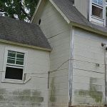 A person in a neon safety vest and face mask stands outside a small, tilted house with white siding, possibly awaiting light demolition or shed removal. The damaged house leans to one side, with trees and a wooden fence in the background.