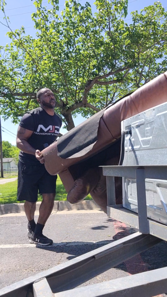 A man wearing a black t-shirt and shorts lifts a brown sofa onto a metal trailer for furniture removal outdoors on a sunny day, with green trees and grass in the background.
