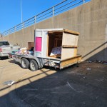A trailer parked near a concrete wall holds a large wooden crate and various items, including boxes and a portable toilet—some debris is scattered nearby. A pickup truck from TX by Arlington Junk Removal Service is parked next to the trailer.
