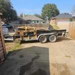 A flatbed trailer loaded with wooden pallets and materials—possibly awaiting a junk removal service—is parked on a driveway in a suburban neighborhood, with houses, trees, and a wooden fence in the background under a clear blue sky.