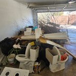 A cluttered garage filled with trash bags, broken toilets, a stove top, cardboard, branches, and various debris awaits a garage cleanout, with the door open to a bright outdoor area.