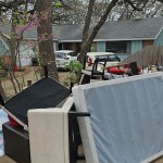 A pile of furniture, likely from an eviction cleanup, including a mattress, chairs, and tables, sits on a sidewalk by the street in front of a suburban house with trees and parked cars visible in the background.