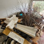 A garage cleanout is needed for this cluttered space, filled with debris like cardboard boxes, Styrofoam pieces, long white panels, leafless branches, buckets, and scattered items. The garage door stands partially open.