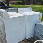 Two white washing machines, one visibly worn and the other in better condition, sit side by side on a wooden surface outdoors—ideal candidates for appliance removal—with grass, trees, and houses visible in the background.