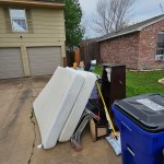 A pile of discarded household items awaits junk removal on a driveway next to blue recycling and black trash bins, in front of a house under a cloudy sky.