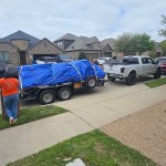 A white pickup truck, likely used for junk removal, is parked on a residential street, towing a trailer covered with a blue tarp. A person in an orange shirt walks nearby as brick houses line the background.