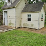 A small, weathered, pale yellow house with two sections sits in a grassy yard. The siding shows signs of mildew and dirt. Windows are on both sections, and a tree with green leaves is behind the house—possible site for light demolition by Arlington Junk Removal Service.