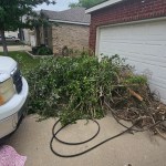 A large pile of tree branches and yard debris is stacked on a driveway in front of a garage at a suburban house, awaiting yard debris removal by Arlington Junk Removal Service. A white vehicle is parked nearby, and a garden hose lies on the ground.